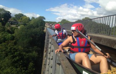 Two individuals in helmets and life jackets canoeing on a bridge over a green landscape.