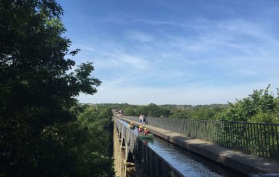 People kayaking on a narrow waterway atop a stone aqueduct under a clear blue sky.