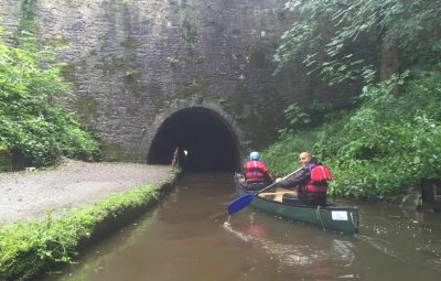 Two individuals in a canoe approaching a dark tunnel on a waterway surrounded by greenery.