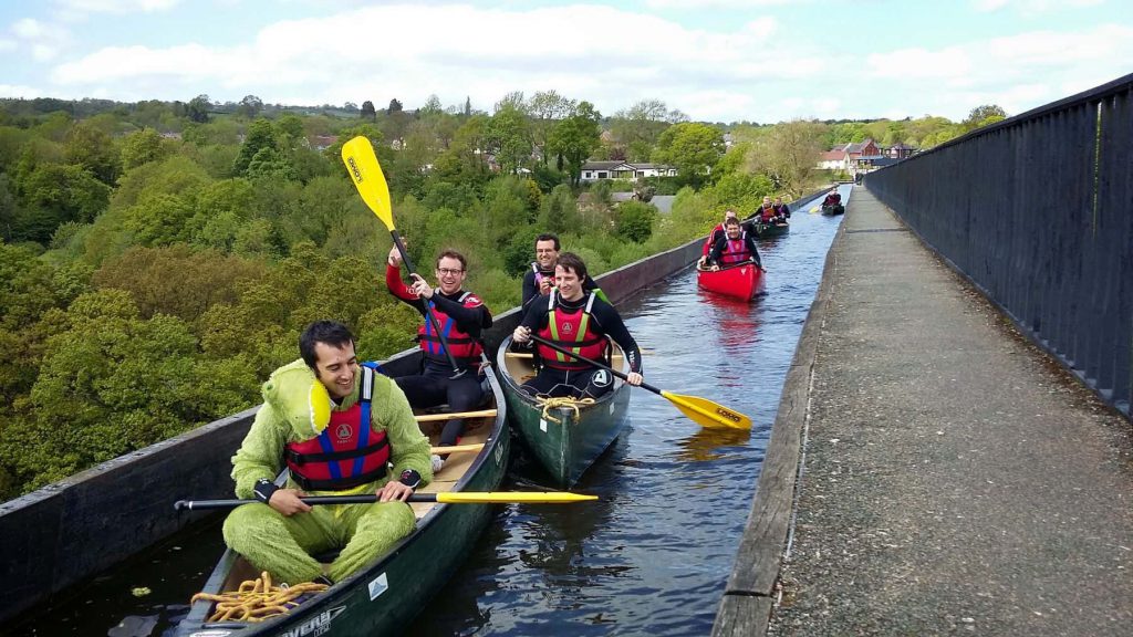 A group of people in canoes paddling on a waterway, some wearing life jackets and holding paddles.