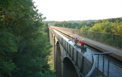 Two kayakers paddling on a waterway beneath a bridge surrounded by greenery.