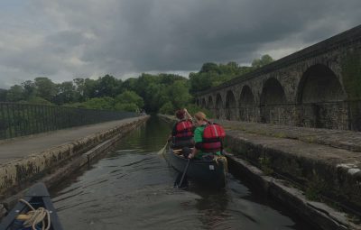 Two individuals in a canoe wearing life jackets paddling along a waterway with stone arches in the background.