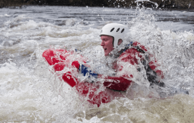 Individual wearing a helmet and red wetsuit bodyboarding through white water waves.