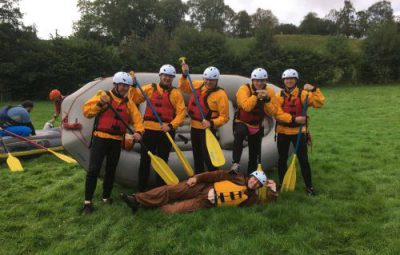 Group of six individuals wearing rafting gear and helmets posing in front of an inflatable raft.